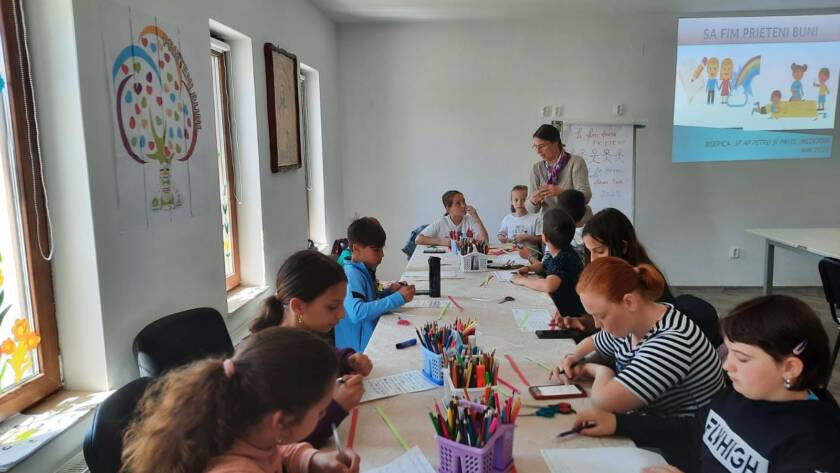Copii lucrând la mesele lungi din sală, cu o prezentare proiectată în fundal