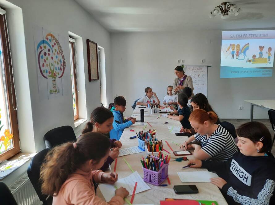 Copii lucrând la mesele lungi din sală, cu o prezentare proiectată în fundal
