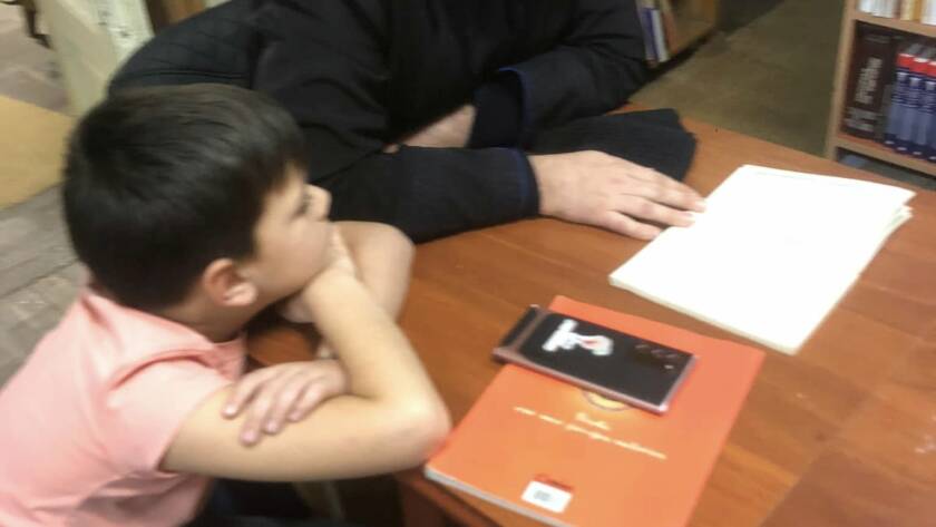Un preot citind dintr-o carte la masă, în fața unui copil atent, în sala bibliotecii