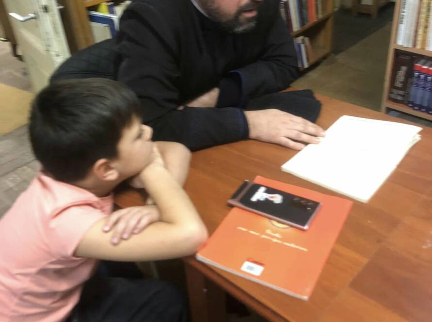 Un preot citind dintr-o carte la masă, în fața unui copil atent, în sala bibliotecii