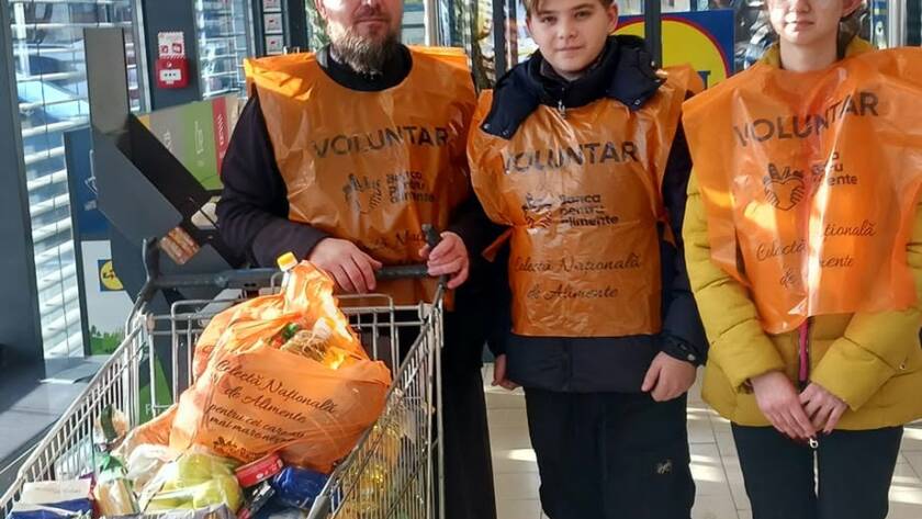 Trei voluntari cu veste portocalii lângă un cărucior cu alimente, la intrarea în magazin