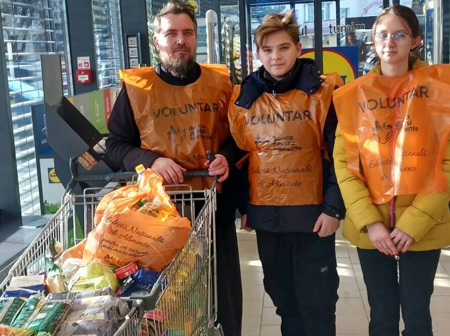 Trei voluntari cu veste portocalii lângă un cărucior cu alimente, la intrarea în magazin