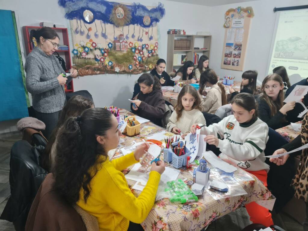 Copii lucrând la machete de hârtie la masă, în sala parohială, în timp ce o doamnă urmărește activitatea