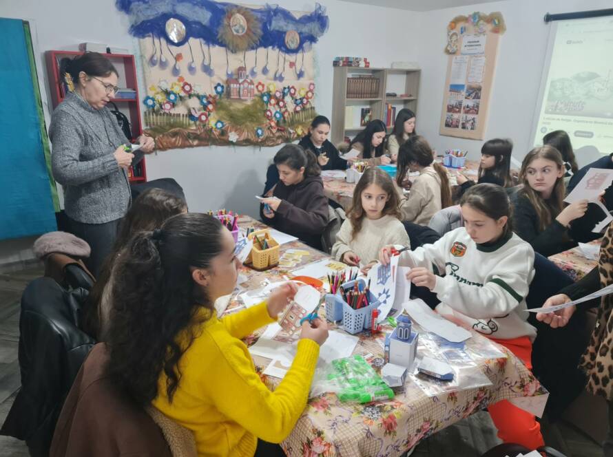 Copii lucrând la machete de hârtie la masă, în sala parohială, în timp ce o doamnă urmărește activitatea